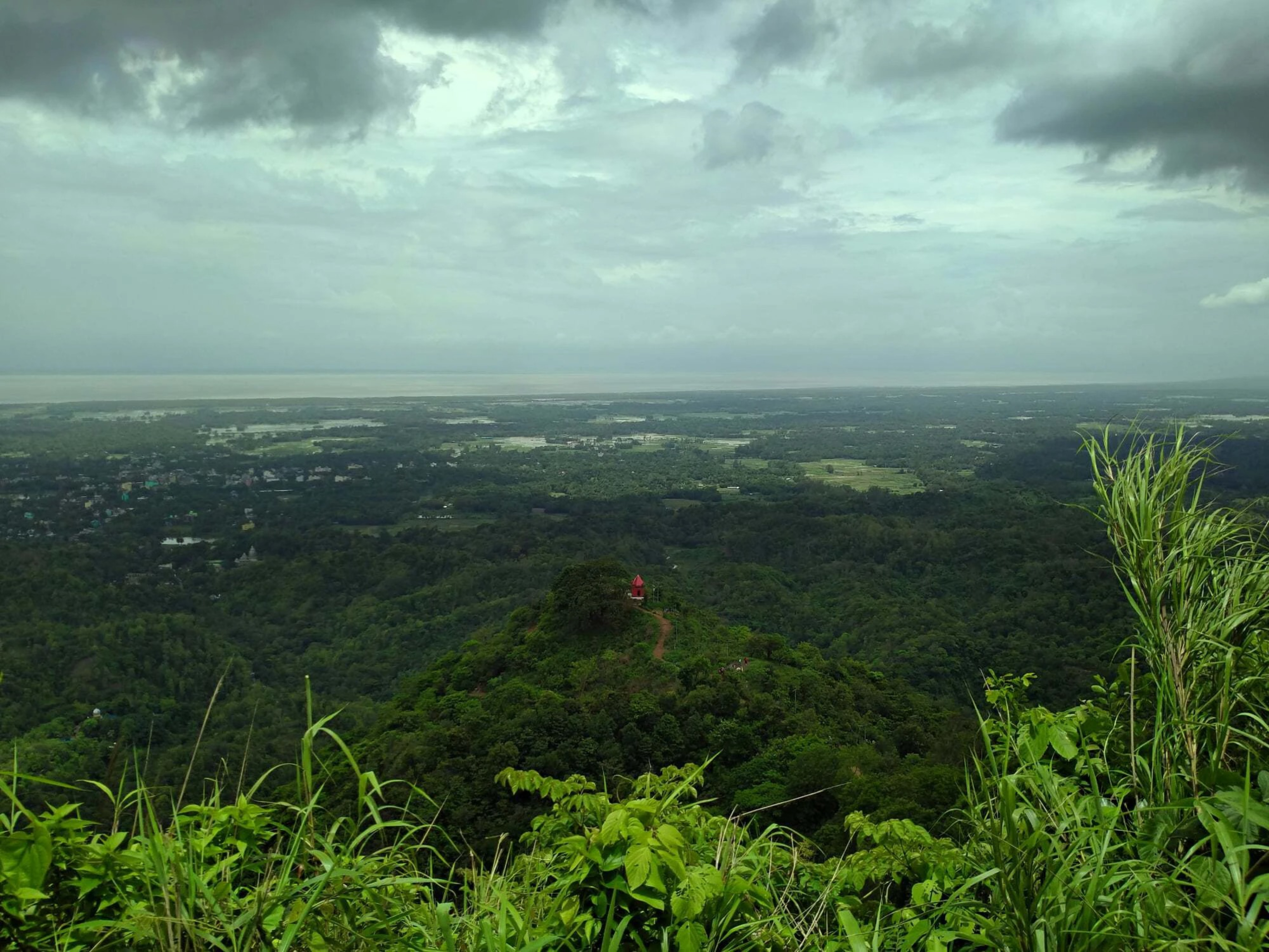 A wide, scenic view of the Chandranath Temple, a small red structure on a forested hilltop in Sitakunda, Bangladesh, under a dramatic cloudy sky