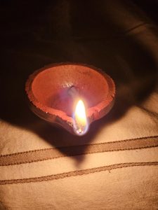 A clay lamp (diya) is glowing warmly in the dark, placed on a cloth-covered surface during a traditional festival night. Captured from Perumanna, Kozhikode.