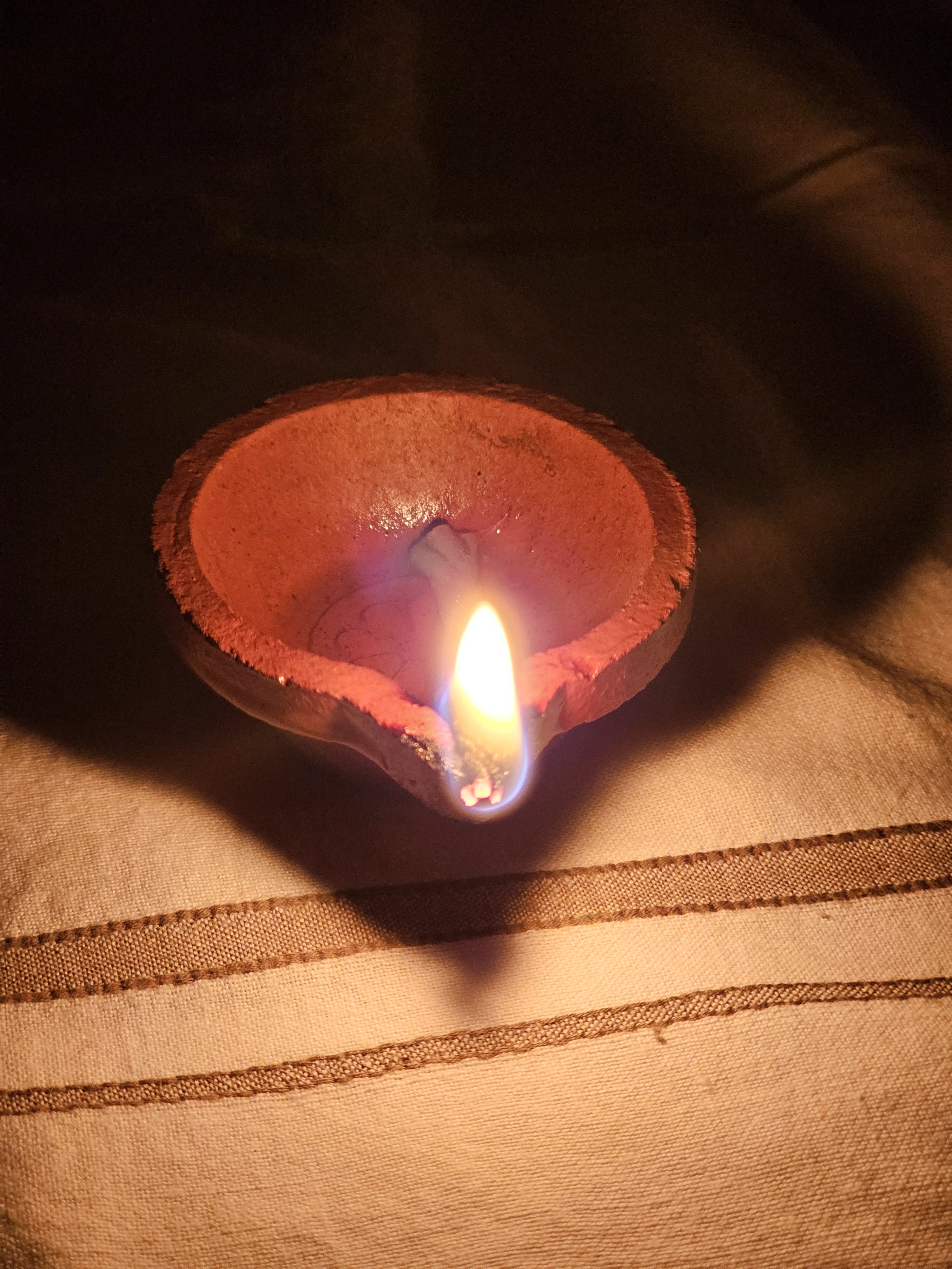 A clay lamp (diya) is glowing warmly in the dark, placed on a cloth-covered surface during a traditional festival night. Captured from Perumanna, Kozhikode.