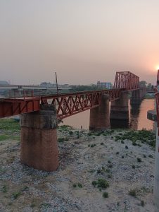 A view of a red railway bridge spanning over a river, supported by large stone pillars
