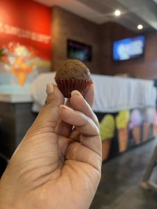 A tiny, dark muffin in a red cupcake liner is held between a person's fingers. 