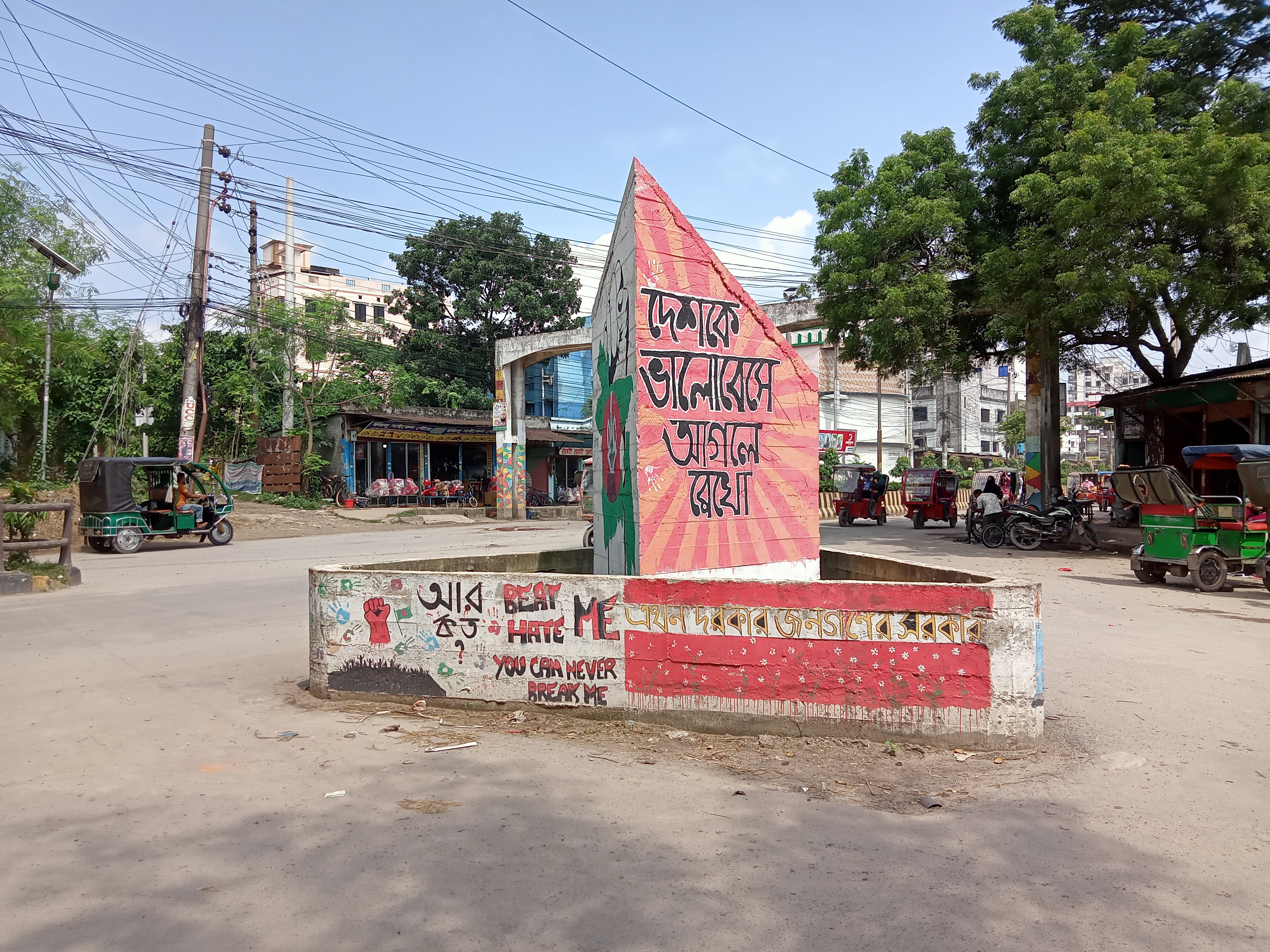 A street corner features a colorful mural on a stone structure with a pyramid-like top, displaying Bengali text. The surrounding area is filled with greenery and utility poles, while a small shop with blue walls is in the background.
