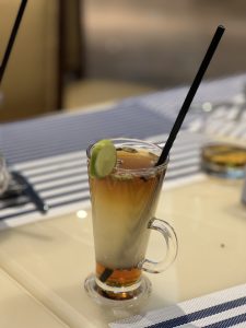A tall, clear glass filled with a layered drink featuring a dark amber top and a lighter bottom, garnished with a slice of lime. The glass has a curved handle and contains a black straw. The drink is placed on a light-colored table with a striped blue and white placemat in the background.