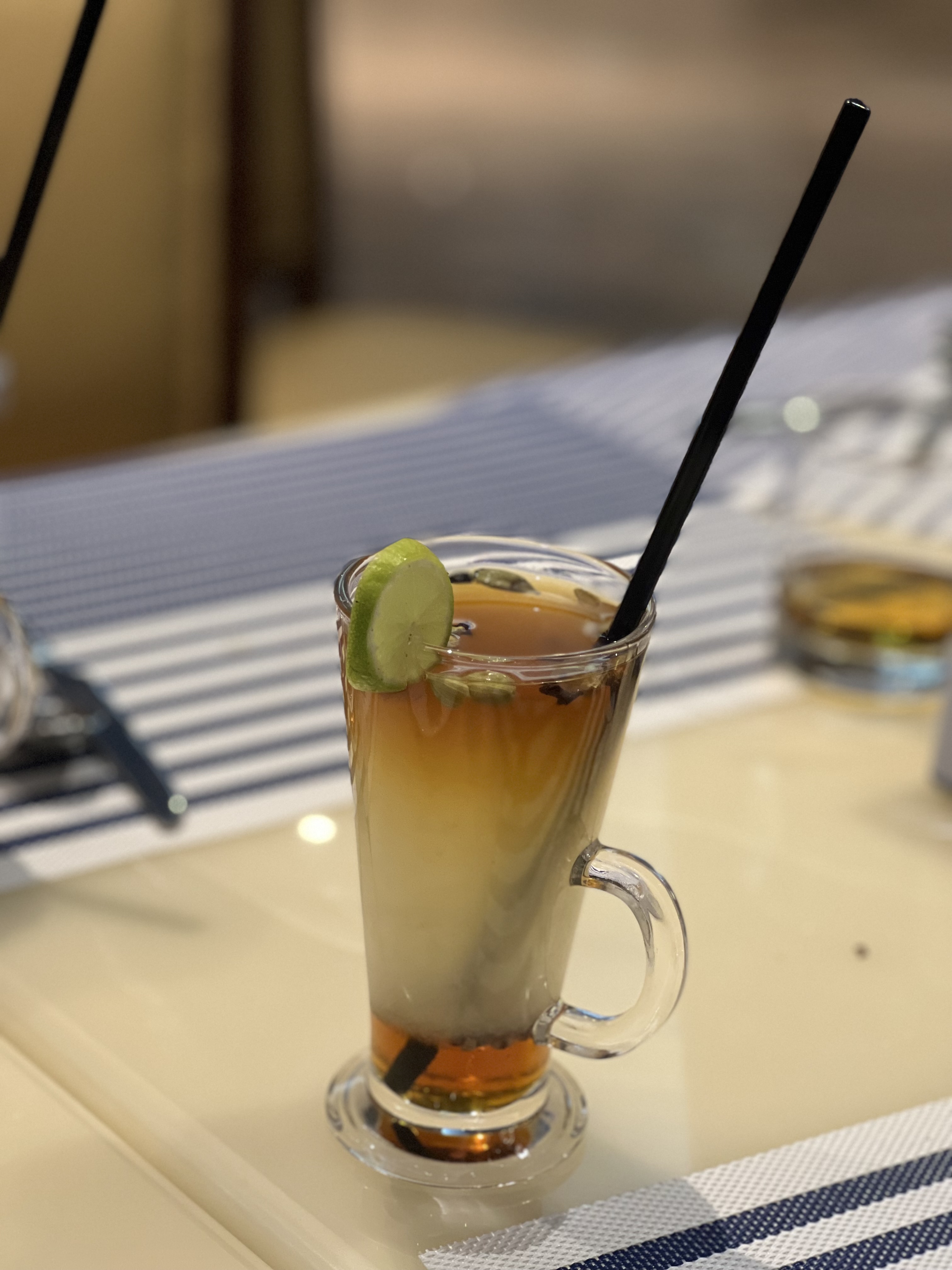 A tall, clear glass filled with a layered drink featuring a dark amber top and a lighter bottom, garnished with a slice of lime. The glass has a curved handle and contains a black straw. The drink is placed on a light-colored table with a striped blue and white placemat in the background.