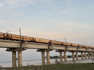 A long yellow and red train crosses a concrete bridge above a calm river at sunset.