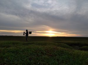 A silhouette of a person walking across a grassy landscape during sunset, holding a stick over their shoulder with bags hanging from it.
