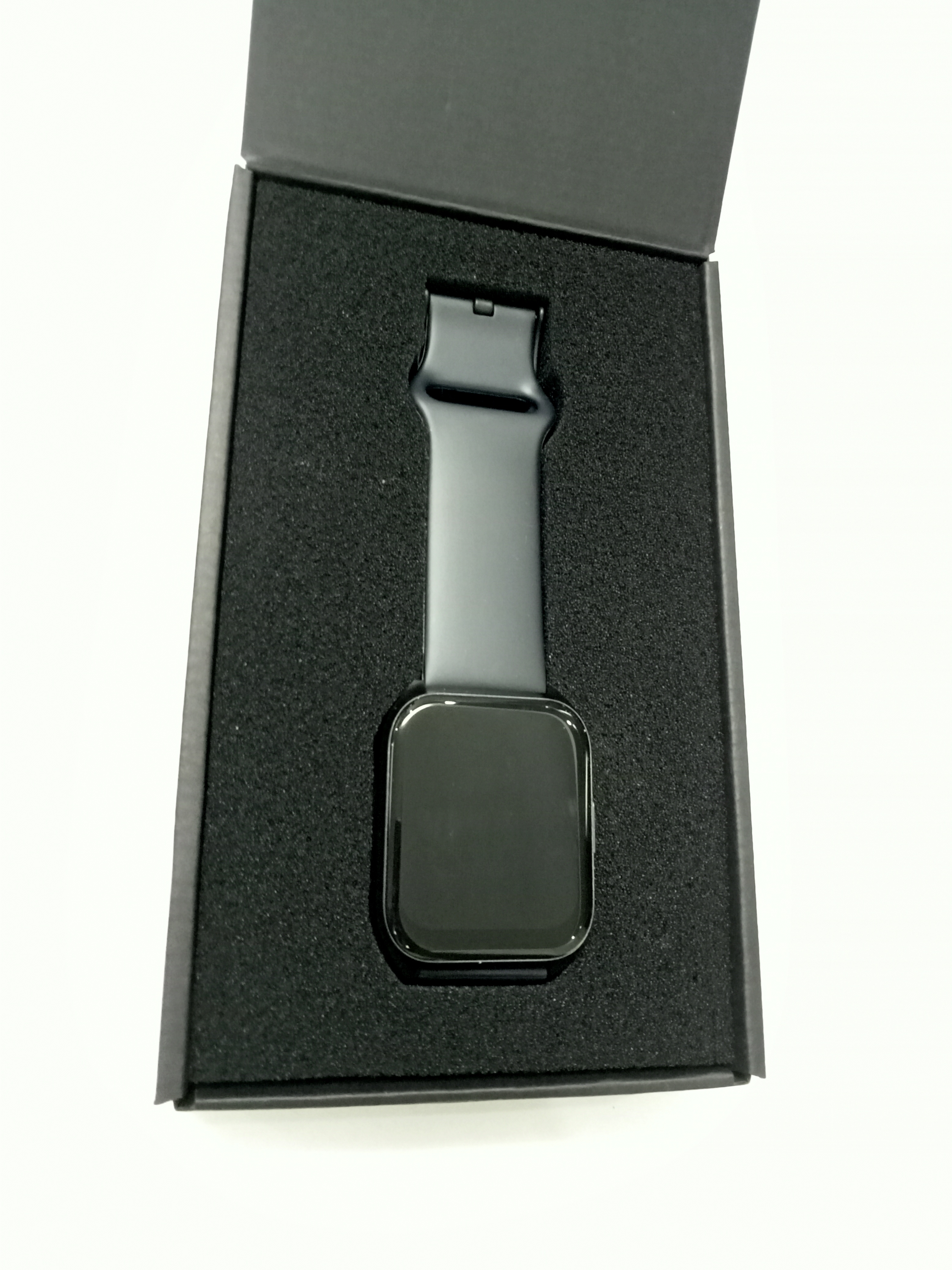 A black smartwatch is resting inside a black cardboard box, which is lined with black foam for protection. The watch features a square face and a black silicone strap, creating a sleek and modern appearance.