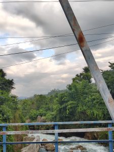 A scenic view from a metal bridge overlooking a river. The foreground features a rusted blue railing and a rocky bank, while the river flows beneath, surrounded by lush greenery.