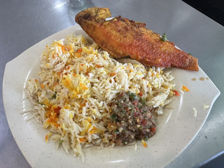 A plate on a metal table with colorful rice, a whole fried fish, and spicy relish.
