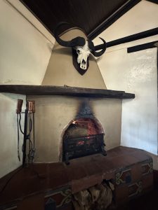 A close-up of a fireplace with a lit fire in a rustic room. Above the mantel, a large skull with long horns is mounted on the wall. A set of metal fireplace tools hangs on the left side of the fireplace, and a pile of firewood rests in a small alcove below the hearth.