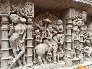 A detailed stone relief sculpture depicting multiple figures in intricate poses. The scene features a woman gracefully standing with a hand to her lips, alongside a figure on horseback, holding a musical instrument, and another figure offering something. The artwork is adorned with ornate columns and elaborate carvings, showcasing artistic craftsmanship and thematic elements of movement and expression. The color of the stone is a warm beige, suggesting historical and cultural significance.