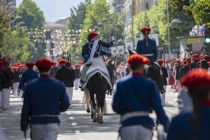 Two riders dressed in traditional Basque military uniforms, wearing red berets and blue jackets, lead the parade on horseback during the Alarde de San Marcial in Irun, Spain, as rows of participants in red berets, black jackets, and white trousers line the street