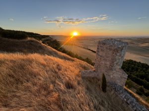 Sunset over rolling fields in Castrojeriz (Burgos, Spain): a weathered stone tower stands on a grassy ridge. Golden light ignites the dry hillside, sunrays streak across the valley, and dark pines dot the slopes as distant plains fade beneath a blue-orange sky.