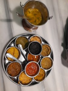 A stainless steel thali with eight colorful Indian dishes, including chutneys and curries, is arranged around a central vibrant red sauce
