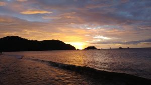 Sunset on the beach with the sun setting behind the mountains and the sea illuminated in shades of gold and orange, while small waves lap at the shore and several boats rest on the horizon
