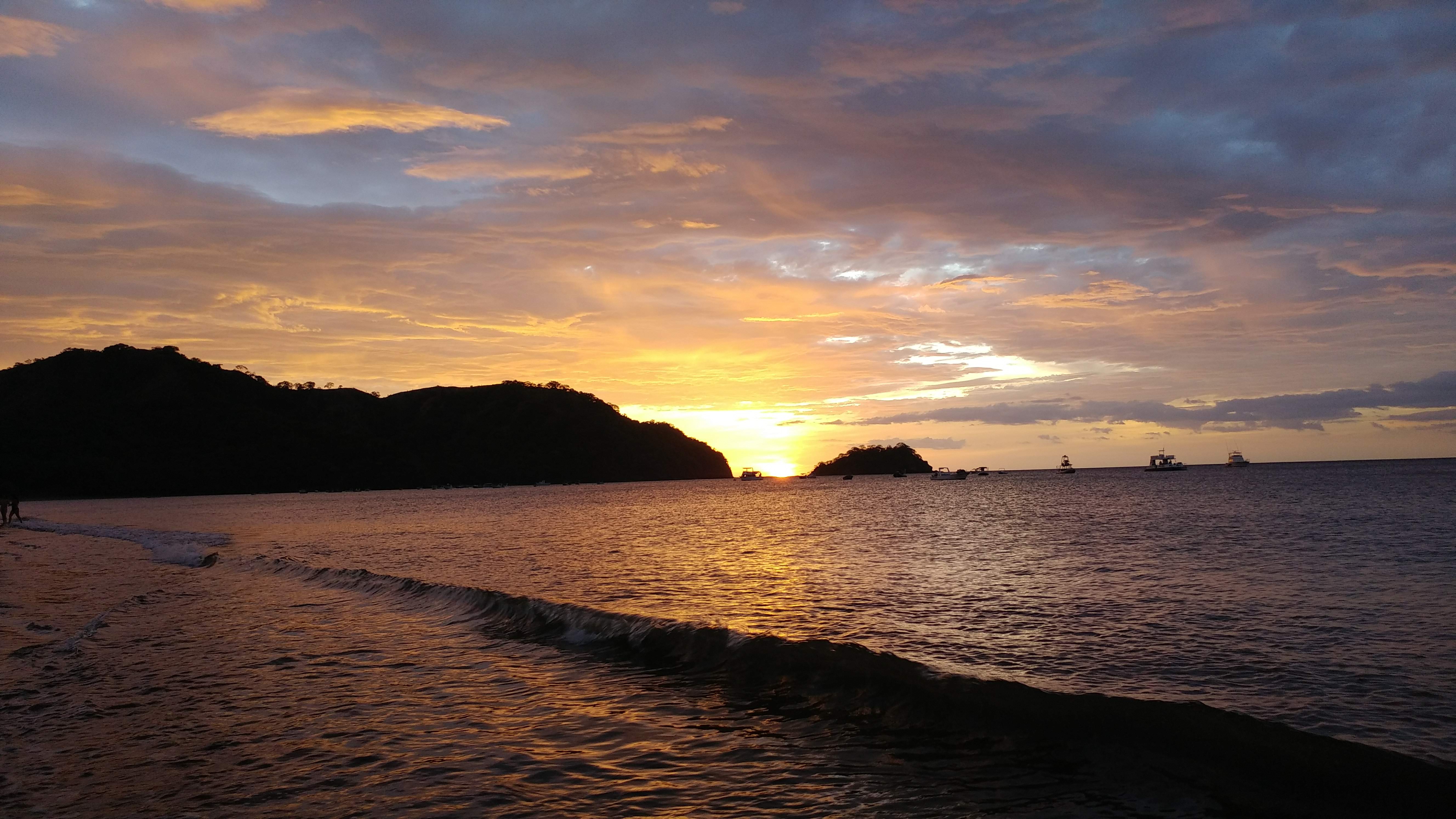 Sunset on the beach with the sun setting behind the mountains and the sea illuminated in shades of gold and orange, while small waves lap at the shore and several boats rest on the horizon