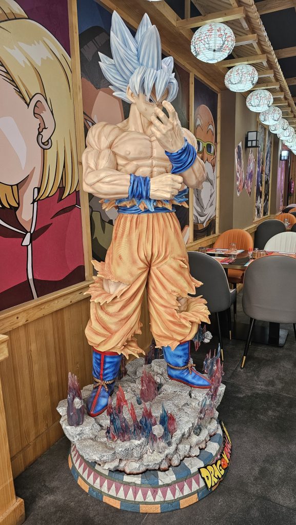 A detailed statue of a spiky blue-haired character in an orange and blue outfit, standing on a rocky base with colorful crystals, set against vibrant murals and decorative lanterns.