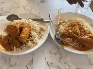 Two plates of white rice topped with a rich, orange-colored chicken sauce are displayed on a marble table. Each plate features pieces of chicken, one with a wing and the other with a drumstick, accompanied by metal utensils. A glass of water is partially visible in the background.
