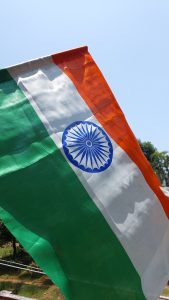 A vibrant Indian flag, featuring its saffron, white, and green stripes, along with the Ashoka Chakra at its center, is depicted waving against a clear blue sky. 
