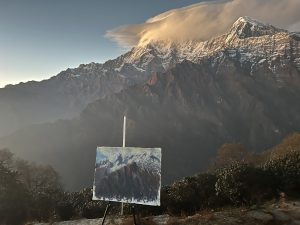 An artist's canvas on an easel sits in the foreground, painting a snow-capped mountain range under a cloud-streaked sky during sunrise