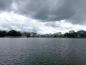 The image presents a large lake under dark cloudy sky with buldings and trees in its background. Dharmasagar Lake, Cumilla, Bangladesh. #SummerPhotoContest