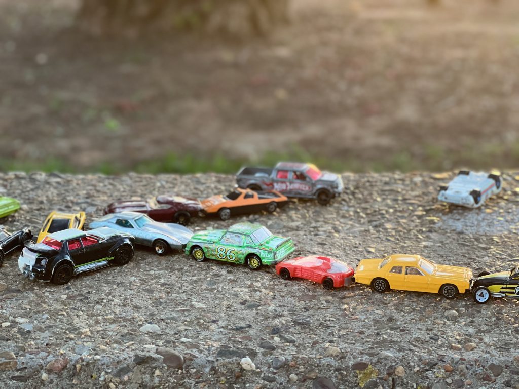 A collection of colorful toy cars is arranged on a gravel surface. The cars are of various styles and colors, including red, yellow, green, and black, creating a playful scene. The background is softly blurred.