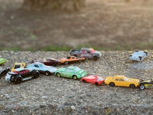 A collection of colorful toy cars is arranged on a gravel surface. The cars are of various styles and colors, including red, yellow, green, and black, creating a playful scene. The background is softly blurred.
