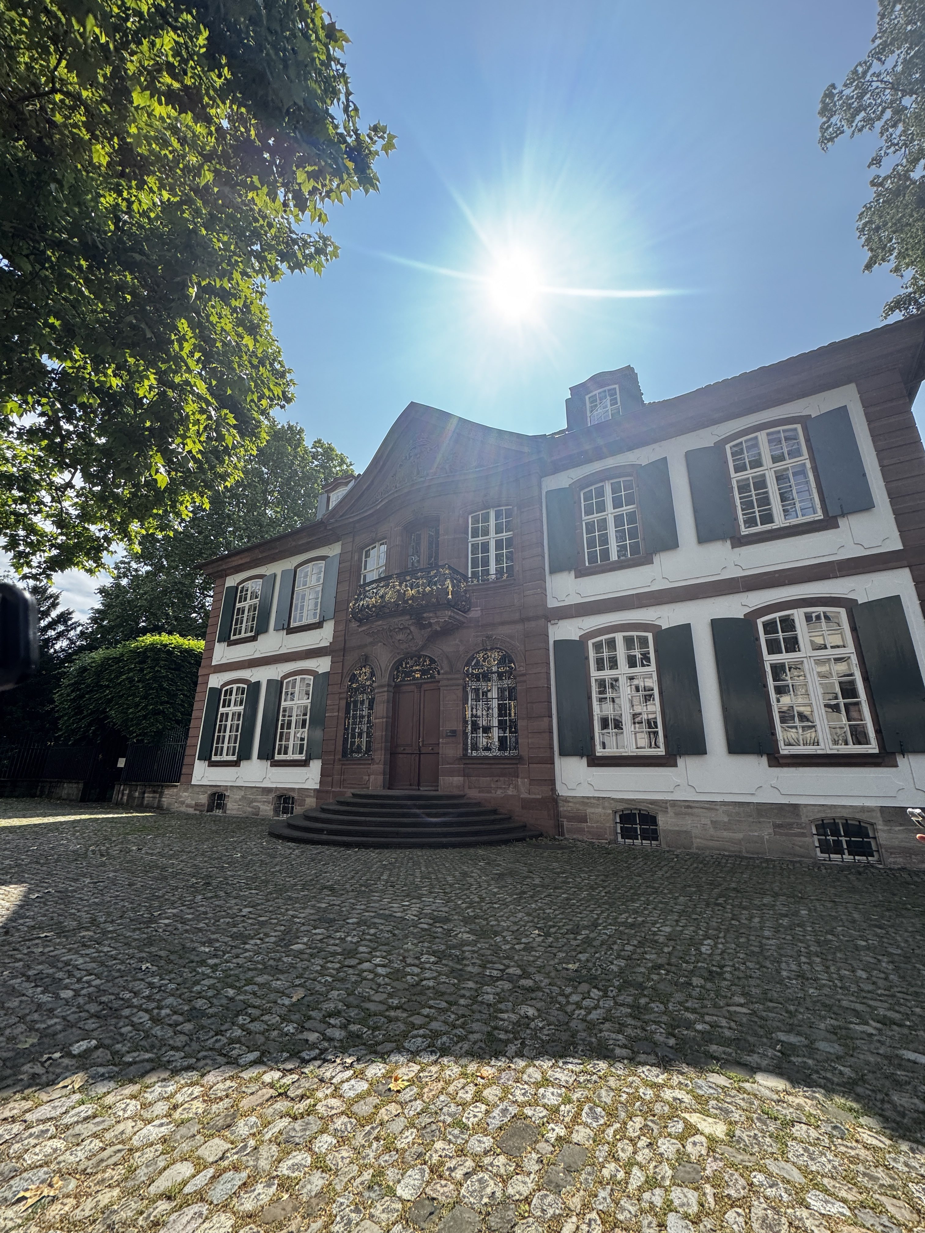 An old building that looks like a large home, with a cobblestone courtyard. The sun is very bright in a clear sky. Basel, edificio antiguo, luz de dia.