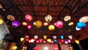 A vibrant collection of colorful, illuminated lanterns hangs from a wooden ceiling, creating a festive atmosphere in Vietnam.