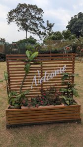 A welcome sign in a garden entrance with several flower pots and trees at its behind. #SummerPhotoContest