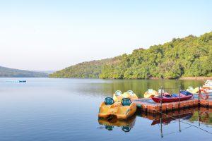 A serene lake surrounded by lush green hills, with a wooden dock featuring yellow pedal boats and a red kayak. The water is calm and reflects the boats and the greenery, under a clear blue sky.