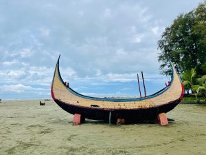 A colorful, traditional-style boat with a high, curved bow and stern rests on a sandy beach under a cloudy sky.