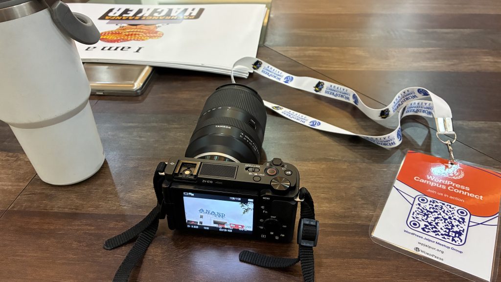 A camera with a Tamron lens is positioned on a wooden table, displaying a captured image on its screen. Nearby, there is a white travel mug, a smartphone, and a badge attached to a lanyard that reads “WordPress Campus Connect” with a QR code. A booklet is partially visible in the background.