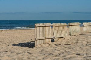 A wooden fence partially buried in soft sand leads toward a tranquil beach scene, with gentle waves lapping at the shore and a clear blue sky overhead.