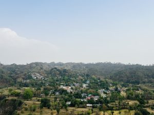 A panoramic view of a lush green landscape featuring rolling hills and a cluster of houses nestled among trees. The houses display a variety of colors, with some roofs in shades of red and blue. Kangra Fort, Himachal Pradesh
