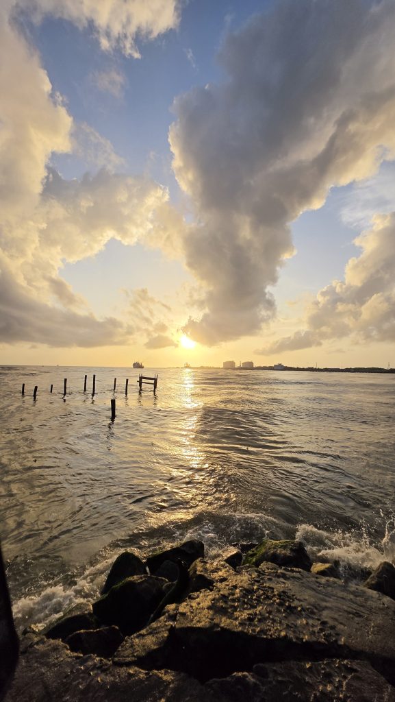 A serene sunset over a sea, with the sun low on the horizon casting a golden reflection on the rippling water. #SummerPhotoContest