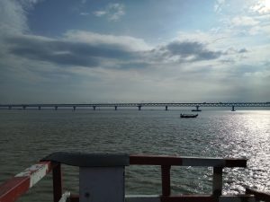 Scenic view of Bangladesh’s Padma Bridge under a partly cloudy sky, with a small boat gliding on the sunlit river in the foreground.