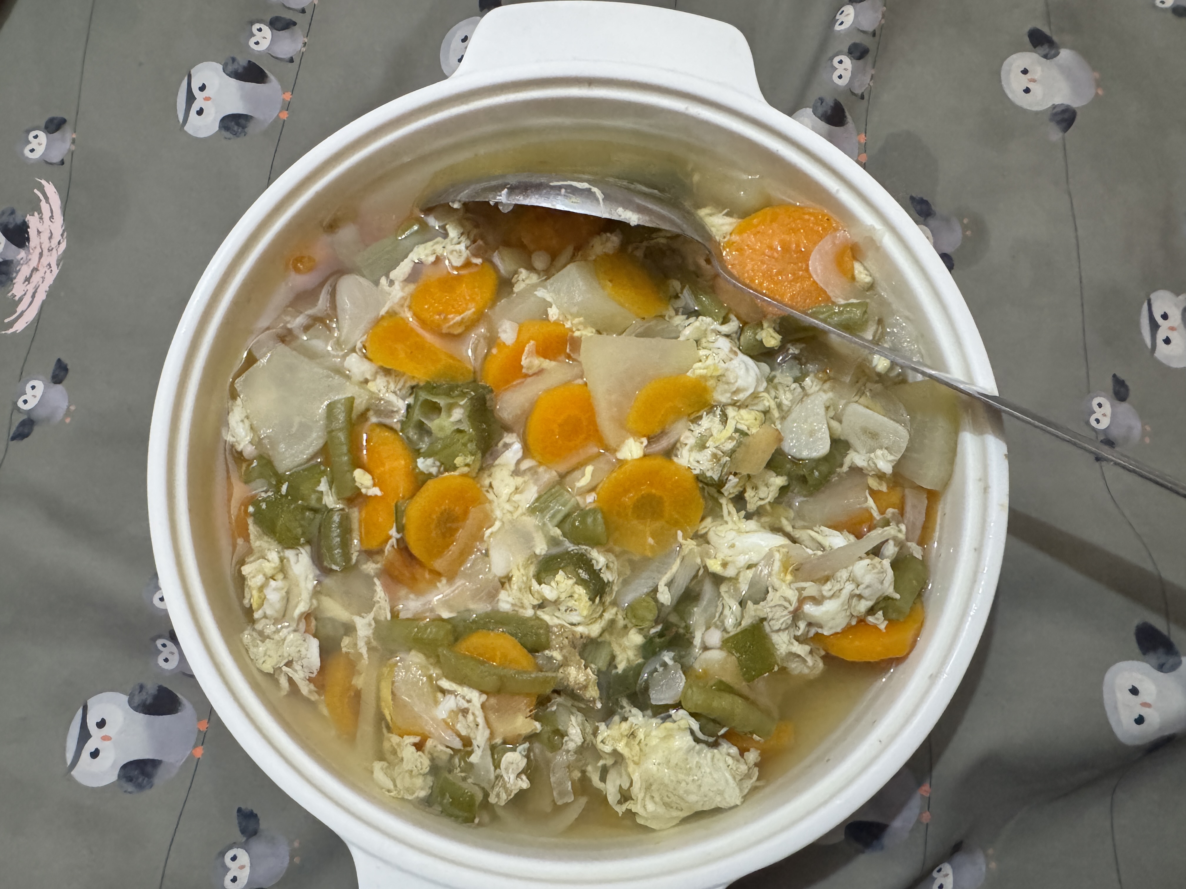 A white bowl of vegetable and egg drop soup sits on a penguin-patterned tablecloth, filled with carrots, green beans, and fluffy egg bits, with a spoon resting inside.