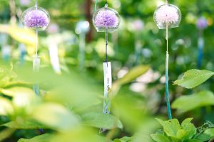 Glass wind chimes with delicate artificial flowers hanging in a lush green garden. A tranquil summer scene that evokes Japanese tradition, serenity, and seasonal beauty.
