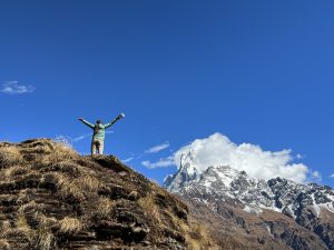 A person stands triumphantly with arms raised on a grassy, rocky ridge. In the background, a majestic, snow-capped mountain with a pointed peak rises under a clear blue sky with a few clouds