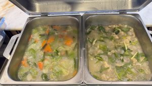 Two buffet trays hold a Thai-style vegetable soup&mdash;greens and carrots in a clear broth on the left, and a chicken or egg version with a slightly cloudy broth on the right.