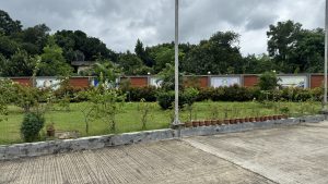 Looking across a wide concrete pavement towards a landscaped garden area under a cloudy sky. The garden features a lawn with small trees, a decorative back wall made of brick and painted panels, and a neat row of small terracotta pots sitting on a ledge, with a dense forest of tall green trees rising up behind