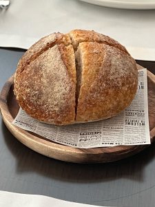 A round loaf of bread with a cross-shaped cut on a wooden plate, resting on a paper with text.
