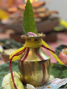 A traditional brass pot adorned with red and yellow fabric, topped with a green leaf and a pink flower, is placed on a bed of rice on banana and tea leaves with a nepali currency 10 rupee. In the background, blurred elements suggest a festive setting with candles or lamps.
