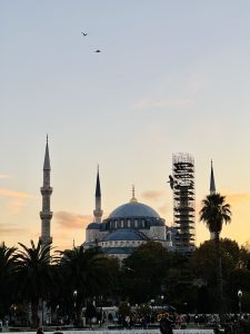 A scenic view of a historical mosque at sunset, featuring minarets and a large dome. The mosque is partially covered with scaffolding, indicating ongoing restoration work. In the foreground, palm trees and people can be seen, while birds fly against a colorful sky with soft hues of orange and blue. The atmosphere is tranquil and picturesque, capturing the essence of a beautiful evening.