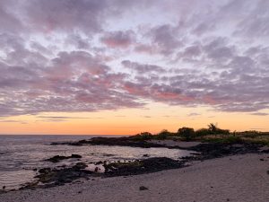 A wide-angle shot of a vibrant orange, pink, and purple sunset over Kailua Beach, Hawaii, with a rocky volcanic shore, sandy beach, and the Pacific Ocean stretching into the distance under cotton candy clouds.
