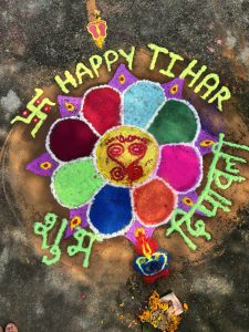 A vibrant rangoli with a central floral circle in red, green, blue, pink, and yellow, surrounded by a lamp symbol, "Happy Tihar" in bright yellow, green text, and scattered floral decorations.