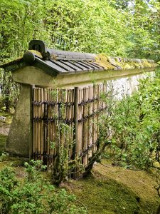 A traditional bamboo and stone wall, featuring a tiled roof partially covered in moss, blends seamlessly into the lush surroundings. Seen at the Portland Japanese Garden. 