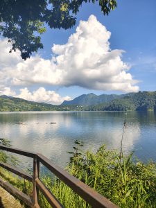 A tranquil lakeside scene featuring calm waters reflecting fluffy white clouds and green hills under a bright blue sky.