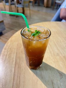 A single glass of iced tea with mint and a green straw sits on a bright wooden cafe table.